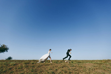 美丽的婚礼 photosession。年轻迷人的新娘在白色花边礼服与长羽跑为她的未婚夫在黑色西装绕着蓝天的背景走在大绿场