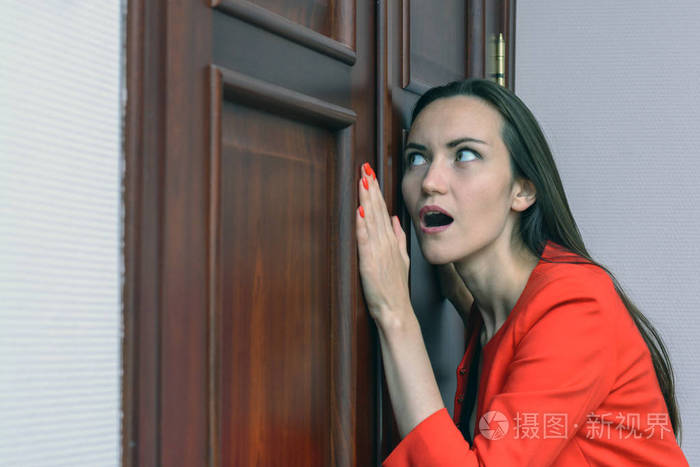 穿红色西装的妇女的水平特写肖像偷听, 窥探老板的门
