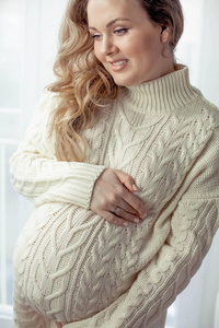 一个年轻漂亮的孕妇站在窗边。穿着舒适的针织礼服怀孕..。母亲.怀孕.一个快乐的孕妇的肖像。舒适