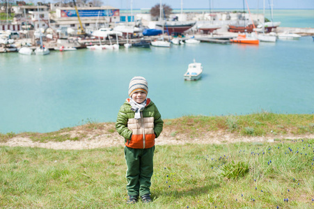 温暖的夹克和帽子和围巾的快乐的小男孩在海湾与船的背景下的绿色斜坡在早春