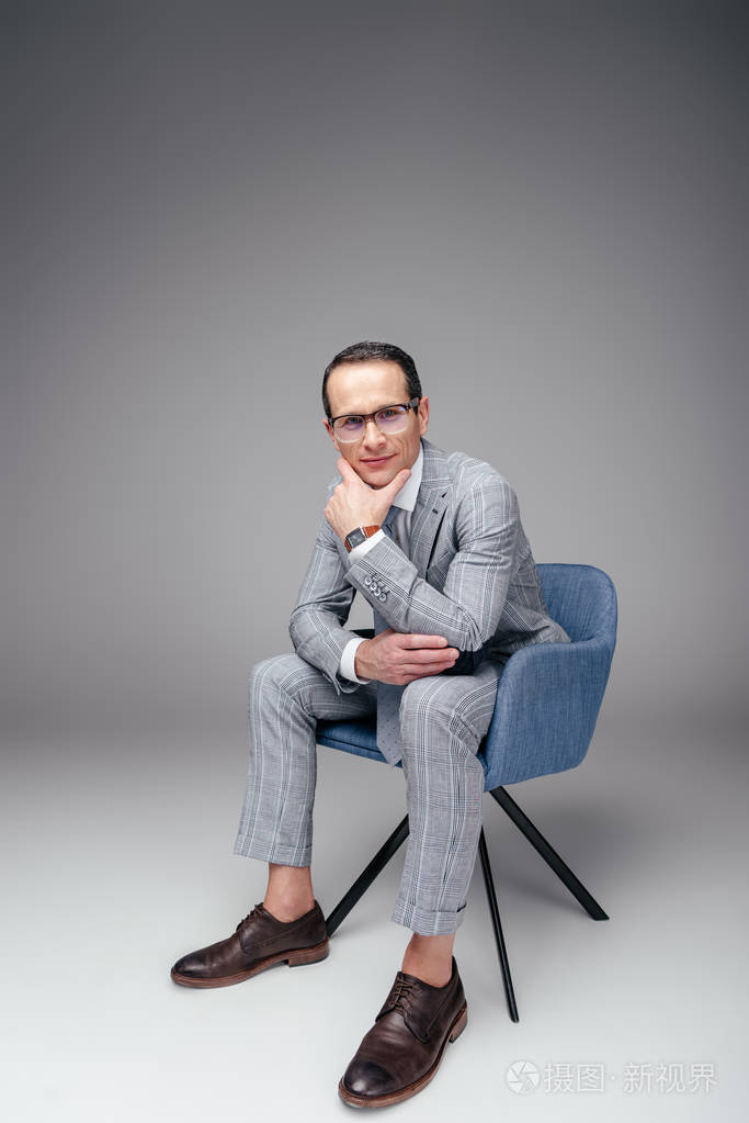 英俊的成年商人穿着时髦西装坐在椅子上, 手放在下巴上灰色