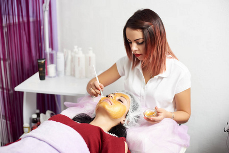 年轻的美容师在一个现代美容院的黑发客户脸上涂上了金色面具