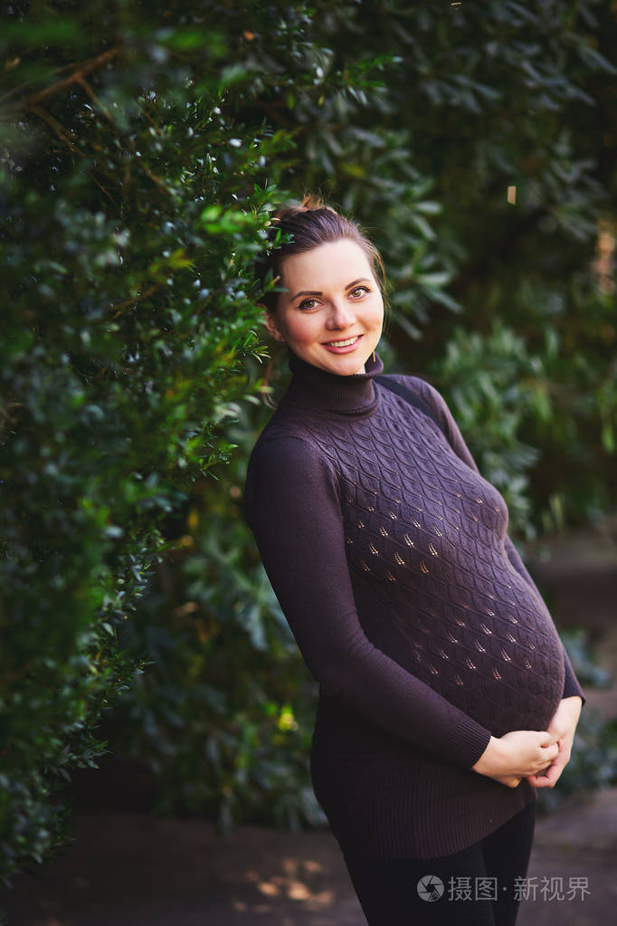 一个微笑着美丽的孕妇的肖像与闪闪发光的外观在针织毛衣抱在她的肚子