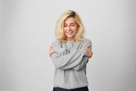 年轻的金发女孩的肖像拥抱自己, 穿着休闲灰色毛衣, 感觉舒适和温暖.