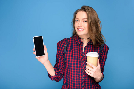 美丽的年轻女孩拿着杯子在咖啡和显示智能手机, 孤立的蓝色