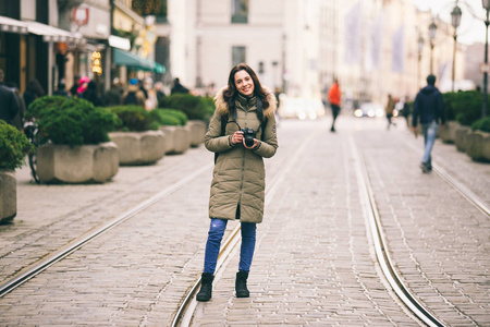 美丽的年轻妇女游人以长的头发在中心街道的背景站立在德国慕尼黑的城市在冬天。持有黑色大的专业相机, 姿势和微笑