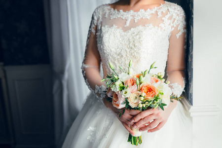 一个美丽的新娘站在窗边, 捧着一束白色玫瑰和桃子牡丹的结婚花束。特写
