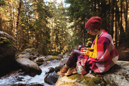 一个漂亮的时髦女孩在她的肩膀上戴着格子格子图案的帽子, 一只黄色的毛衣和眼镜, 手里拿着一杯咖啡, 看着一条山上的河, 坐在松树