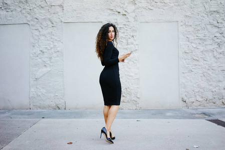 年轻美丽的女人与长卷曲的头发穿着黑色礼服使用她的智能手机对白色墙壁背景