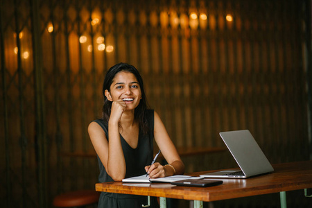 有吸引力和年轻的印度 亚洲 专业人士的坦率的肖像在她的笔记本电脑在一个舒适的咖啡馆或 coworking 空间。她附近有笔记