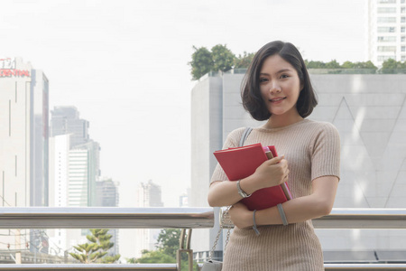 亚洲少女肖像在户外公共空间与现代建筑背景下举行红色书立场和礼服在街道时尚布料