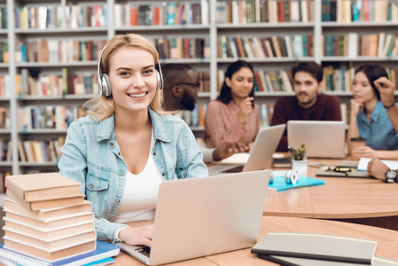 一群多文化的学生坐在图书馆的桌子上, 白人女孩在笔记本电脑上使用耳机