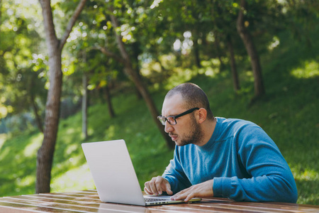 年轻成功的精明男子商人或学生在休闲蓝衬衫眼镜坐在桌子上的手机在城市公园使用便携式电脑户外工作的绿色性质。移动办公概念