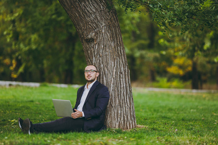年轻成功的商人在白衬衫, 经典西装, 眼镜。男人坐在草地上, 在城市公园的笔记本电脑上工作, 在户外的绿色草坪上自然。移动办公