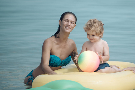 可爱的儿童男孩与坐在空气床垫菠萝形状, 在海洋, 海。母性观念。母亲站在附近, 用儿子推床垫。带着笑脸的妈妈和孩子在海里共度时光