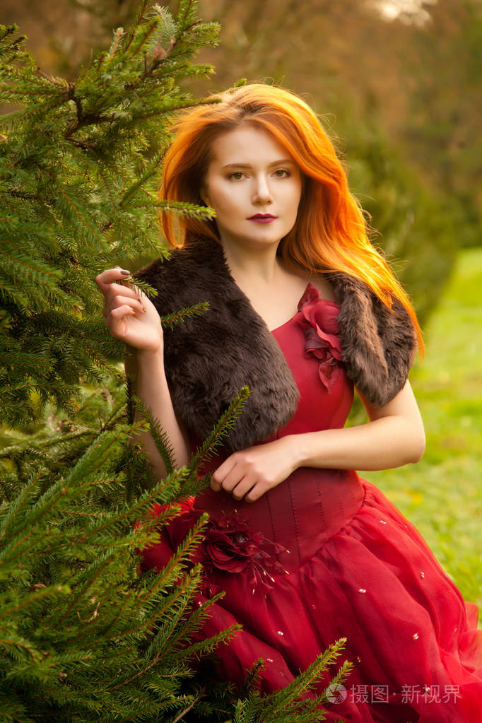 美丽的年轻红发女孩穿着长晚上的红色礼服, 独自漫步在冷杉树公园