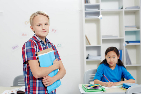 可爱的小学生与抄写本旁看着相机在教室的背景下, 他的同学