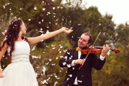 美丽的新娘和新郎玩羽毛在公园与新郎在小提琴演奏