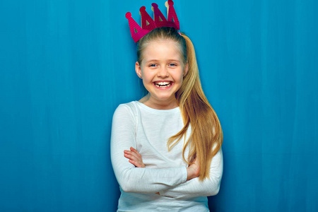 微笑的小女孩与纸冠站立与交叉的胳膊在蓝色墙壁背景