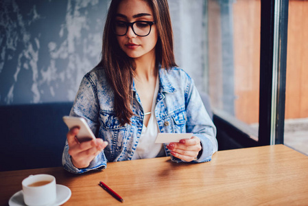 严肃的年轻妇女在时尚眼镜持有名片在手和拨打手机号码在现代智能手机。成功的女性打字手机信息在咖啡馆坐在桌上
