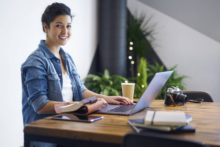 微笑成功的博客的肖像坐在桌子上的现代笔记本电脑和工作时, 看着相机。时尚 coworking 空间上网本设备中的快乐女性研究