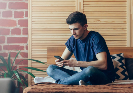 后视图的时髦手忙着用手机, 年轻的男学生打字时坐在床上专心于手