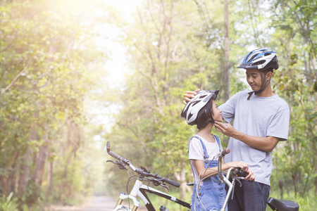 快乐的父亲和女儿骑自行车在公园给他的女儿戴着脚踏车头盔。
