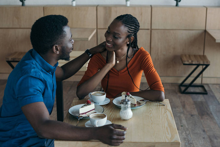 愉快的非洲美国人夫妇坐在桌与杯子咖啡和甜点在咖啡馆