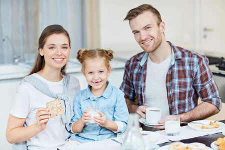 微笑可爱的女孩与牛奶玻璃和她的父母坐在餐桌上吃早饭