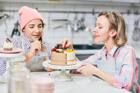 两个年轻的妇女装饰美味的节日蛋糕与异国情调的水果和浆果