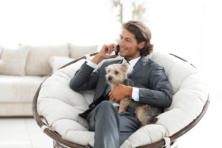 商人握着他的宠物，会谈在智能手机上，而坐在舒适的椅子上