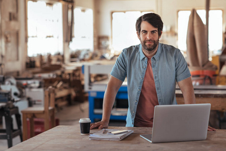 一个自信的年轻的木工胡子的肖像, 在他的大型车间, 靠在工作台上, 用笔记本电脑在线工作