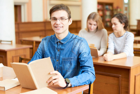 在大学图书馆坐在办公桌前准备期末考试时, 英俊的大学生穿着牛仔衬衫摆出牙齿微笑的腰上画像