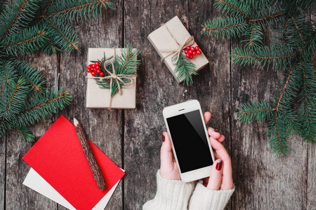 女手握住手机上木制背景与圣诞礼物，冷杉枝。圣诞节和新年快乐组成。平躺，顶视图