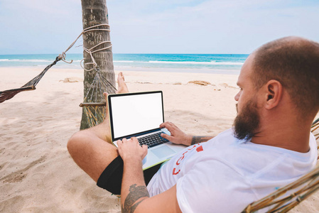 男商人游客在电脑键盘上键入文字, 空白屏幕为您的互联网网站在热带岛屿上休息。年轻人自由职业者工作在膝上型电脑在距离工作躺在吊床上