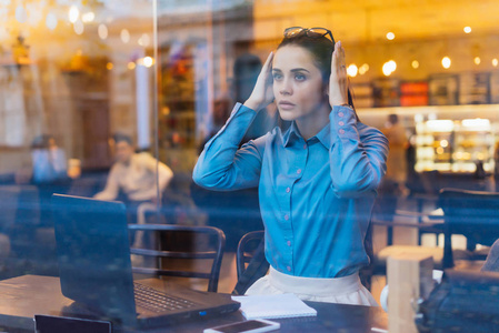 疲惫沉思年轻的自由职业者女孩坐在咖啡馆, 在笔记本电脑, 看着窗外