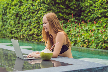 年轻女性自由职业者坐在游泳池附近与她的笔记本电脑。假期很忙。遥远的工作概念