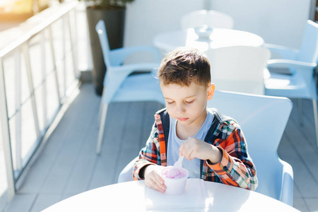 可爱的男孩儿童在格子衬衫吃草莓冰淇淋在一个茶杯里坐在一个咖啡馆在夏日的街道上