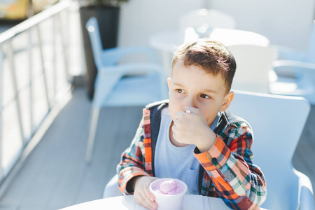 可爱的男孩儿童在格子衬衫吃草莓冰淇淋在一个茶杯里坐在一个咖啡馆在夏日的街道上