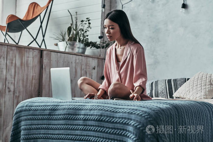 严肃的亚洲年轻妇女使用电脑笔记本电脑和写笔记, 而坐在家里的床上