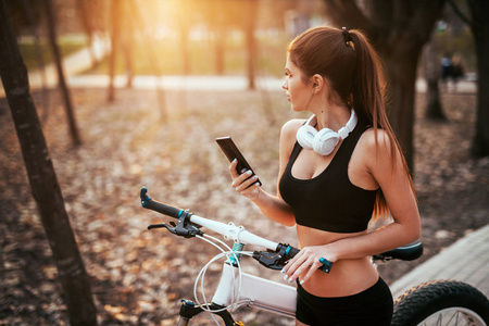 体育女孩与耳机站立与电话和看在距离在自行车附近在公园的日落