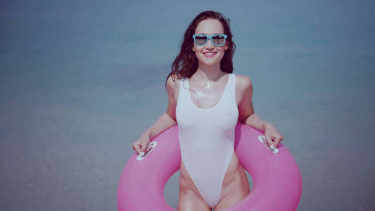 美丽快乐的年轻女子在泳装和太阳镜与粉红色充气环有乐趣在海水中, 享受阳光明媚的夏日在她的海滩热带假日