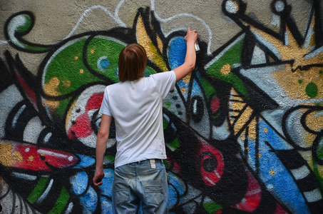 在旧的混凝土墙上画涂鸦图案的过程中的照片。年轻的长发金发男人画了一幅不同颜色的抽象图画。街头艺术与破坏概念