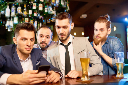 一群惊讶的大胡子男子聚集在酒吧, 喝啤酒和阅读新闻的帮助下的智能手机