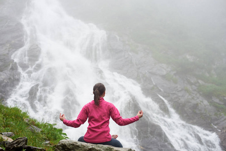 关注在罗马尼亚壮观的 Balea 瀑布前面的岩石练习瑜伽的年轻女性。女人诱人的健康生活方式亥母惊人的惊险景观自然游客