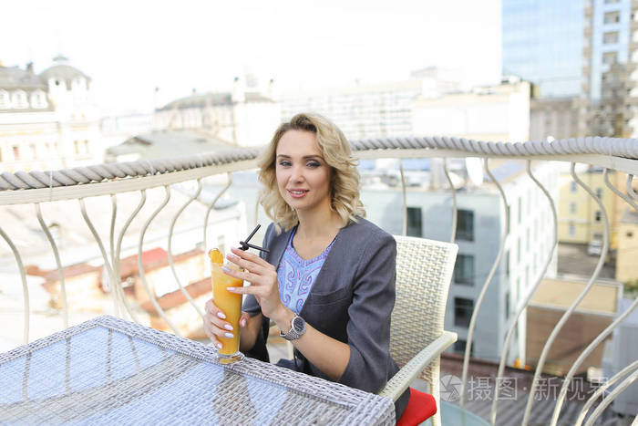 快乐的女人喝果汁在阳台上的咖啡馆与城市背景