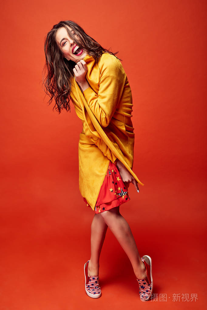 波浪状的头发身着红色的明星印刷礼服和黄色大衣在工作室与深橙色背景