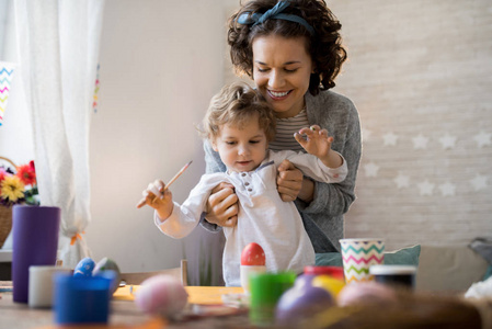 快乐的年轻母亲的肖像帮助小学步儿童油漆复活节彩蛋, 同时准备在家度假