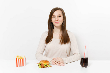 美丽的年轻女子坐在餐桌上, 汉堡, 炸薯条, 可乐在玻璃瓶子上被隔离在白色的背景。适当的营养或美国经典的快餐食品。带有复制空间的
