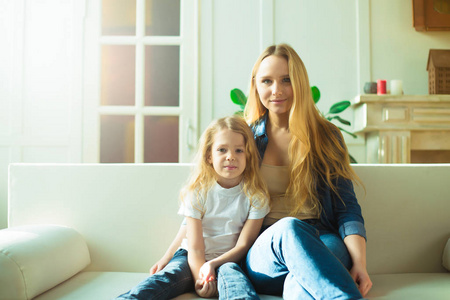 美丽的微笑金发碧眼的母亲和女儿拥抱和在沙发上的乐趣在家里和看相机。母亲和女儿的概念